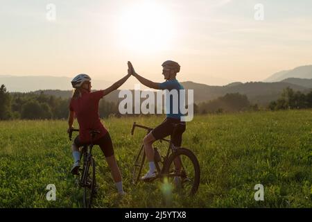 Straßenradfahrer, Mann und Frau, halten an einem grasbewachsenen Hügel, um den Rennerfolg mit hohen fünf Gesten bei Sonnenuntergang zu feiern. Stockfoto