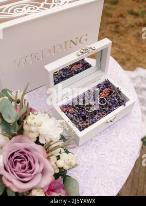 Lavendelhochzeit. Hochzeitsringe mit violetten Blumendekoren auf weißem Tisch in Nahaufnahme. Stockfoto