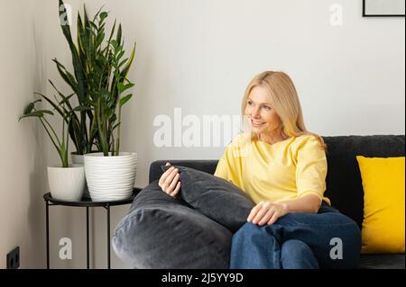 Bezaubernde, sorglose kaukasische Frau, die auf der Couch sitzt und sich entspannt. Porträt einer charmanten älteren blonden Frau, die zu Hause träumt Stockfoto