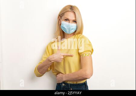 Gesundheitswesen und Medizin. Reife Frau, die eine Schutzmaske trägt, schützt sich mit einem Impfstoff gegen Covid und andere Viruserkrankungen, glückliche ältere Frau zeigt mit dem Finger auf einen medizinischen Fleck auf ihrer Armschulter Stockfoto