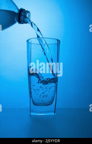 Ökologisch sauberes Wasser wird aus einer Flasche in ein Glas auf blauem Hintergrund gegossen Stockfoto