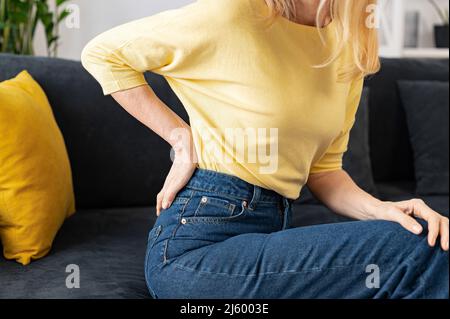 Beschnittene Ansicht der älteren Frau fühlte einen starken Schmerz in Lenden, die zu Hause auf dem Sofa saßen, und besorgte kaukasische Dame, die eine Hand auf dem unteren Rücken hielt Stockfoto