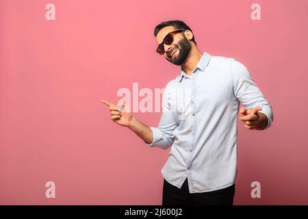 Überglücklich indischer Mann trägt Sonnenbrillen, tanzt, macht Bewegungen zur Musik, lächelt positiv, ist in Hochstimmung. Sorgloser hispanischer Kerl in blauem Jeans-Hemd tanzt isoliert auf Rosa Stockfoto