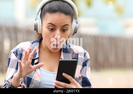 Vorderansicht Porträt eines staunenden Teenagers, der Musik hört und Telefoninhalte in einem Park findet Stockfoto