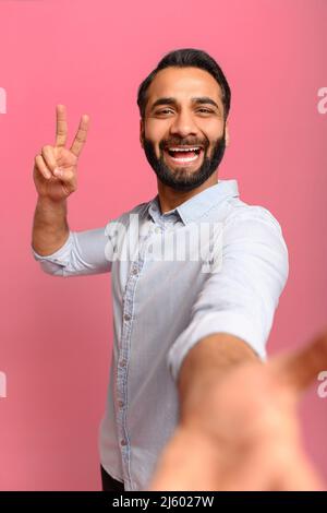 Porträt eines hübschen Mannes aus dem Alter zwischen den Indianern, der Selfie macht, die Kamera anschaut und eine V-Geste mit der Hand zeigt, aus der Perspektive des Fotos. Innenaufnahme des Studios isoliert auf rosafarbenem Hintergrund, vertikal Stockfoto