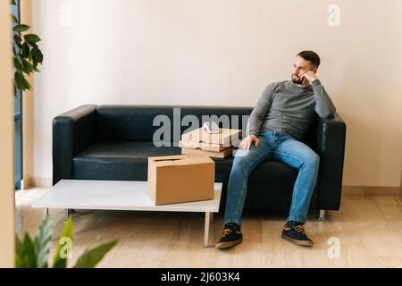 Vorderansicht des Lieferers, der den Kunden wartet, während er auf dem Sofa mit Kartons, Pizza, Pappkarton und dem drahtlosen Zahlungs-POS-Terminal in der Halle sitzt. Stockfoto