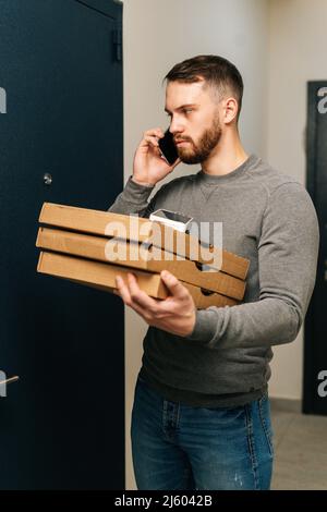 Vertikale Aufnahme des Lieferers, der vor der Tür der Kundenwohnung mit Kartons mit heißer Pizza stand und den Kunden telefonisch anrief. Stockfoto