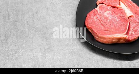 High-Angle-Studioaufnahme eines rohen Rinderrippe-Eye-Steakes auf einer schwarzen Designplatte, die auf einer grauen Betonoberfläche mit freiem Kopierraum steht. Keto-Diät Stockfoto