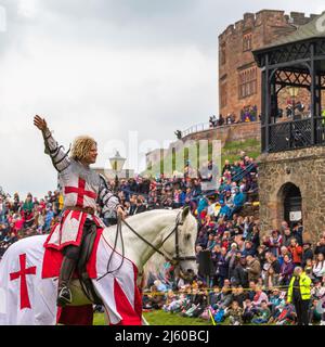Ein Ritter mit Rüstung, der auf einem weißen Pferd sitzt, hebt seine Hand, um bei einer mittelalterlichen Nachstellung der Menge zu winken. Stockfoto