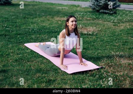 Lächelnde junge Frau, die im Park Yoga macht. Schöne Frau in Sportkleidung liegt in Cobra-Pose und macht Bhujangasana-Übung Stockfoto