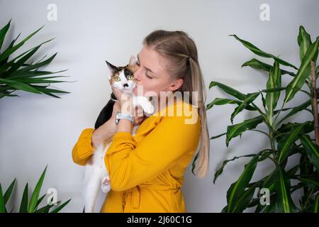 Junge Frau mit ihrer Calico-Katze umarmt und küsst das Kätzchen, das darüber unzufrieden ist Stockfoto