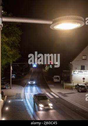 Heiningen, Deutschland. 26. April 2022. Autos fahren unter einer Straßenlaterne, deren Helligkeit durch künstliche Intelligenz gesteuert wird. Nachts soll die Beleuchtung an der örtlichen Durchgangsstraße an das Verkehrsaufkommen angepasst werden und so Insekten schützen. Die Tiere werden vom Licht angezogen und sterben in den Leuchten oder kreisen vor ihnen, bis sie erschöpft sind. Ein Kamerasystem wird auch verwendet, um die Insekten zu zählen. Quelle: Stefan Puchner/dpa/Alamy Live News Stockfoto