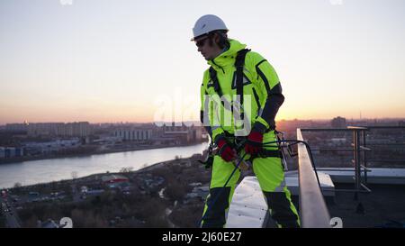 Industriekletterer in einem Sicherheitsgurt und einem weißen Helm wirft ein Seil, während er bei Sonnenuntergang in Zeitlupe auf dem Rand eines Dachs steht Stockfoto