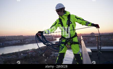 Industriekletterer in einem Sicherheitsgurt und einem weißen Helm wirft ein Seil, während er bei Sonnenuntergang in Zeitlupe auf dem Rand eines Dachs steht Stockfoto