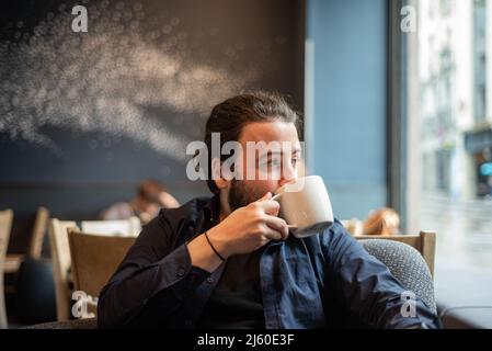 Lange Haare Hispanic junge Erwachsene männlich trinken in einem Becher im Hipster Coffee Shop Stockfoto