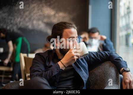 Lange Haare Hispanic junge Erwachsene männlich trinken in einem Becher im Hipster Coffee Shop Stockfoto