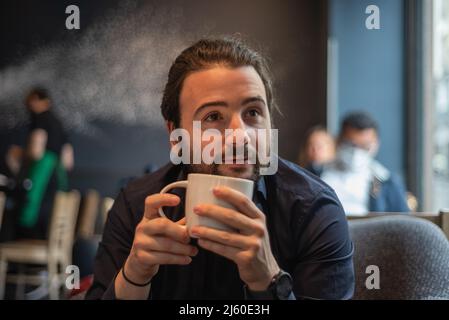 Lange Haare Hispanic junge Erwachsene männlich trinken in einem Becher im Hipster Coffee Shop Stockfoto