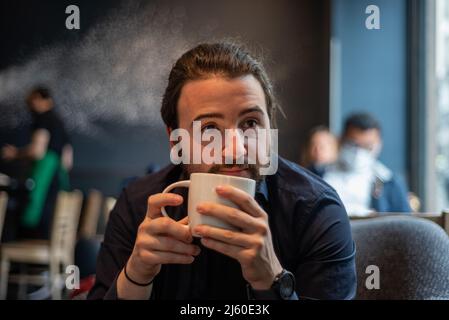 Lange Haare Hispanic junge Erwachsene männlich trinken in einem Becher im Hipster Coffee Shop Stockfoto