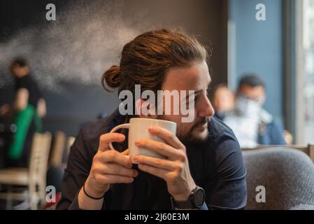 Lange Haare Hispanic junge Erwachsene männlich trinken in einem Becher im Hipster Coffee Shop Stockfoto