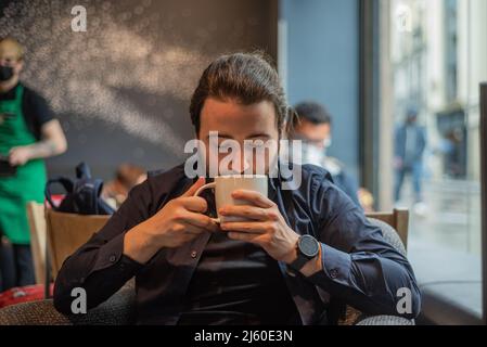 Lange Haare Hispanic junge Erwachsene männlich trinken in einem Becher im Hipster Coffee Shop Stockfoto