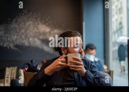 Lange Haare Hispanic junge Erwachsene männlich trinken in einem Becher im Hipster Coffee Shop Stockfoto