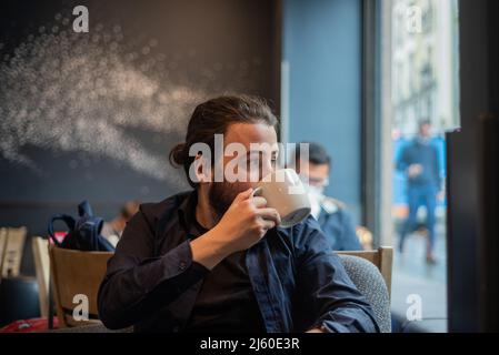 Lange Haare Hispanic junge Erwachsene männlich trinken in einem Becher im Hipster Coffee Shop Stockfoto