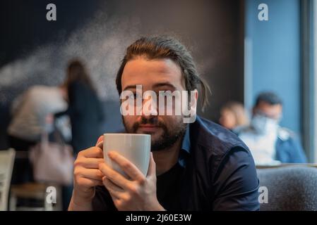 Lange Haare Hispanic junge Erwachsene männlich trinken in einem Becher im Hipster Coffee Shop Stockfoto