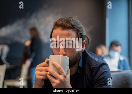 Lange Haare Hispanic junge Erwachsene männlich trinken in einem Becher im Hipster Coffee Shop Stockfoto