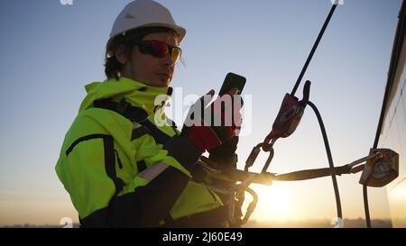 Mann ein Bauingenieur in einem Hut hängt mit einem Telefon in einem Gurt an Seilen an einem hohen Haus und führt bei Sonnenuntergang eine geplante Inspektion durch Stockfoto