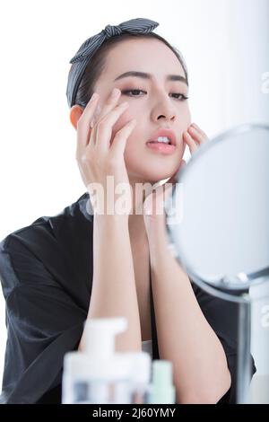 Schöne junge chinesische Frau mit Hautpflegeprodukten vor einem Kosmetikspiegel, auf weißem Hintergrund - Stock Foto Stockfoto