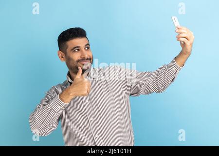 Zufriedener Mann Blogger zeigt Daumen nach oben wie Geste und winken Blick auf Handy-Kamera, machen Selfie oder Video-Aufnahme, trägt gestreiftes Hemd. Innenaufnahme des Studios isoliert auf blauem Hintergrund. Stockfoto