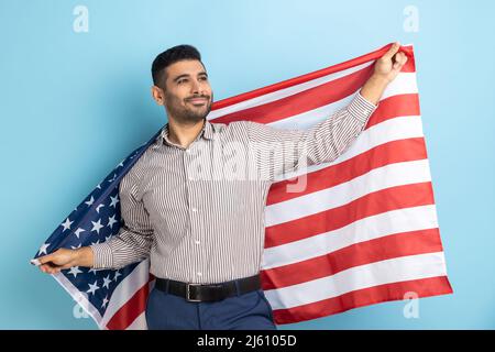 Mann, der die USA-Flagge in den Händen hält, selbstbewusst wegschaut, stolz auf seine soziale Sicherheit im Land, sich sicher fühlt, gestreiftes Hemd trägt. Innenaufnahme des Studios isoliert auf blauem Hintergrund. Stockfoto