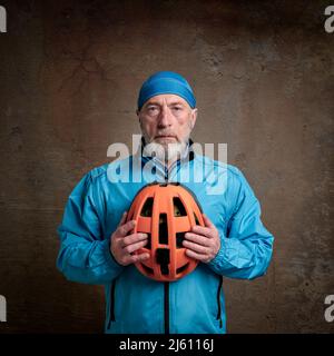 Porträt eines älteren Radfahrers in hellblauer Fahrradjacke und Totenkopf-Mütze, der einen Helm gegen eine grunge Betonwand hält Stockfoto