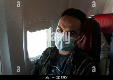 Ein Mann mit Gesichtsmaske, während er in einem Flugzeug sitzt. Neues normales Reisen während einer Pandemie. Männlicher Passagier auf Reisen. Tragen einer Maske in der Flugzeugkabine Stockfoto