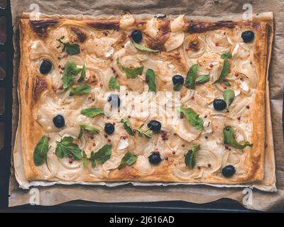 Pizza aus Blätterteig mit Zwiebel, Käse, Oliven, rosa Pfeffer und Feta-Käse oder Schafskäse auf rustikalem Backpapier - Draufsicht, sauberes Essen Stockfoto