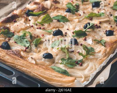 Hausgemachte knusprige Pizza auf Blätterteig-Basis mit Zwiebel, Käse, Oliven, rosa Pfeffer und Feta-Käse oder Schafskäse auf rustikalem Backpapier Stockfoto