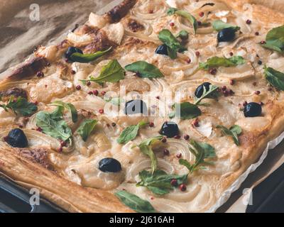 Hausgemachte Pizza auf Blätterteig - fertig zum Essen mit Zwiebel, Käse, Oliven, rosa Pfeffer und Feta-Käse oder Schafskäse auf rustikalem Backpapier Stockfoto