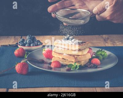 Wie man traditionelle hausgemachte Waffeln mit Puderzucker pulvert - Holztisch ist mit frisch gebackenen handgemachten flauschigen Waffeln dekoriert ein schöner Teller, pl Stockfoto