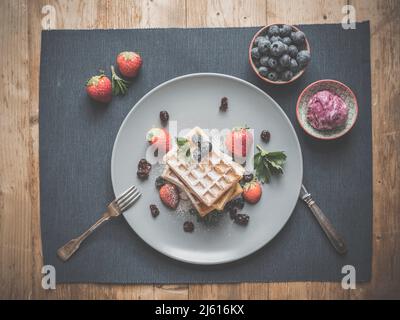 topview von Heidelbeer-Waffeln auf blauem Teller und Textil dekoriert mit mehr Heidelbeeren, Silberbesteck, hausgemachtem Himbeer-Eis und Minze auf Rusti Stockfoto