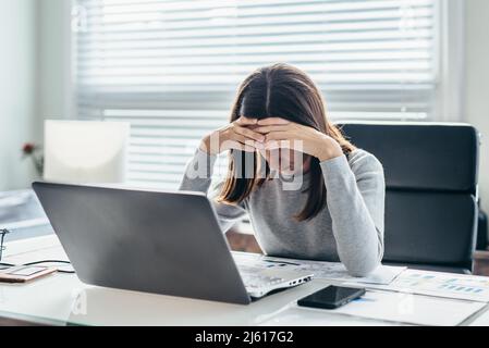 Frau, die im Büro am Tisch saß und ihren Kopf mit den Augen nach unten auf ihre Hand lehnte Stockfoto