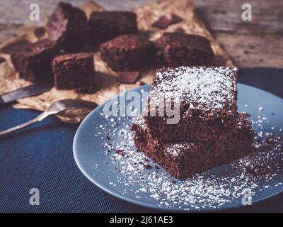 Das Bild zeigt, wie man die besten Brownies macht; rustikaler Holztisch ist mit rustikalem Tischset und einigen weiteren Brownies auf Backpapier dekoriert Stockfoto