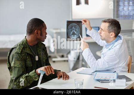 Professioneller Arzt, der während der Verabredung im Krankenhaus einem jungen erwachsenen Militäroffizier ein Röntgenbild des Schädels zeigt Stockfoto