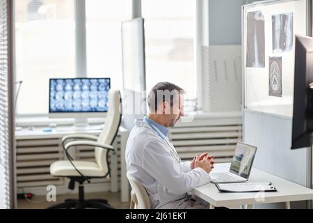 Ein reifer kaukasischer Radiologe mit weißem Labormantel sitzt am Schreibtisch vor dem Laptop und arbeitet mit Tomographieaufnahmen Stockfoto