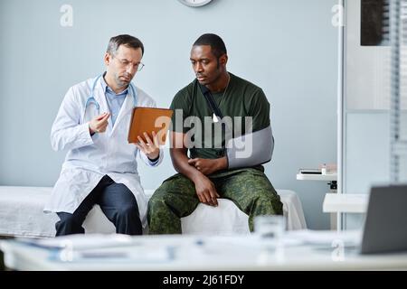 Junger schwarzer Soldat mit gebrochenem Arm und Tragetuch, der auf der medizinischen Couch sitzt und einen Arzttermin hat Stockfoto