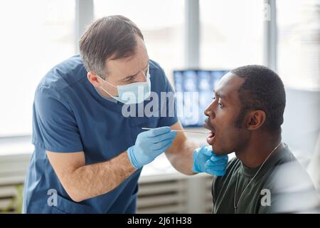 Medizinisches Personal mit Schutzmaske und Handschuhen, die Mundtupfer von jungen schwarzen Soldaten für den PCR-Test nehmen Stockfoto