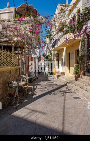 Aegina Island Griechenland - 4.27.2022: Straße mit Geschäften, wandelte ältere Frau mit Schutzmaske in einer engen Stadtallee. Geschlossen Geschäfte, Sommertag Stockfoto