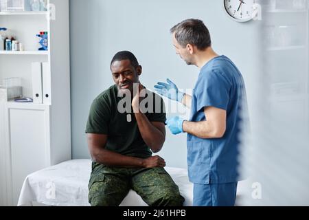 Junger schwarzer Soldat, der im modernen Militärkrankenhaus auf einer medizinischen Couch sitzt und dem Arzt von Nackenschmerzen erzählt Stockfoto
