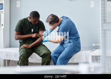 Professioneller Traumatologe, der Schutzhandschuhe trägt und den verletzten Arm eines jungen afroamerikanischen Soldaten antastend Stockfoto