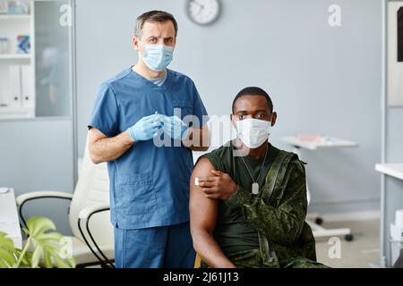 Porträt eines reifen männlichen Krankenpflegers, der die Spritze neben einem jungen schwarzen Militäroffizier hält, der nach einer Covid-Impfung die Kamera anschaut Stockfoto