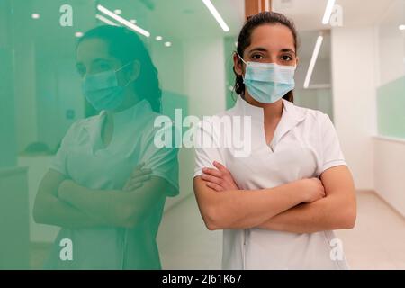 Krankenschwester trägt eine schützende Gesichtsmaske und steht mit den Armen, die von einer Glaswand durchzogen sind, im Flur des Krankenhauses Stockfoto